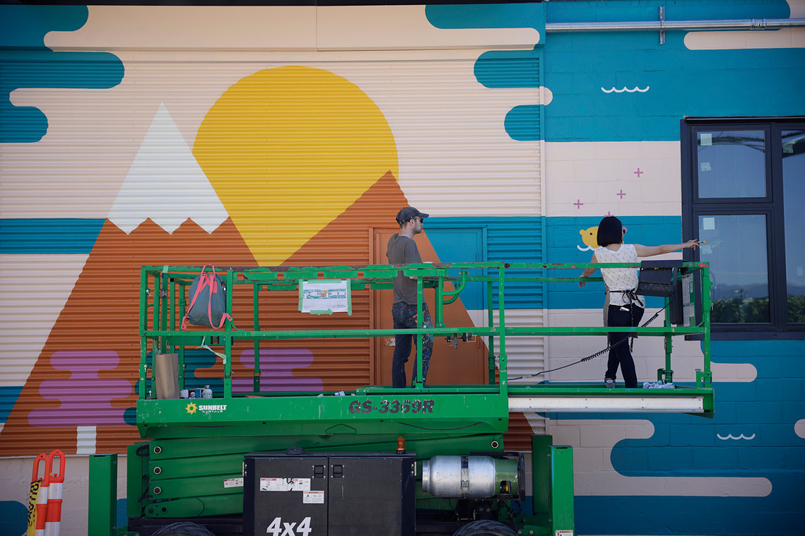 A photo of artist Annie Chen and her assistant Mark Illing working on a mural on a lift.