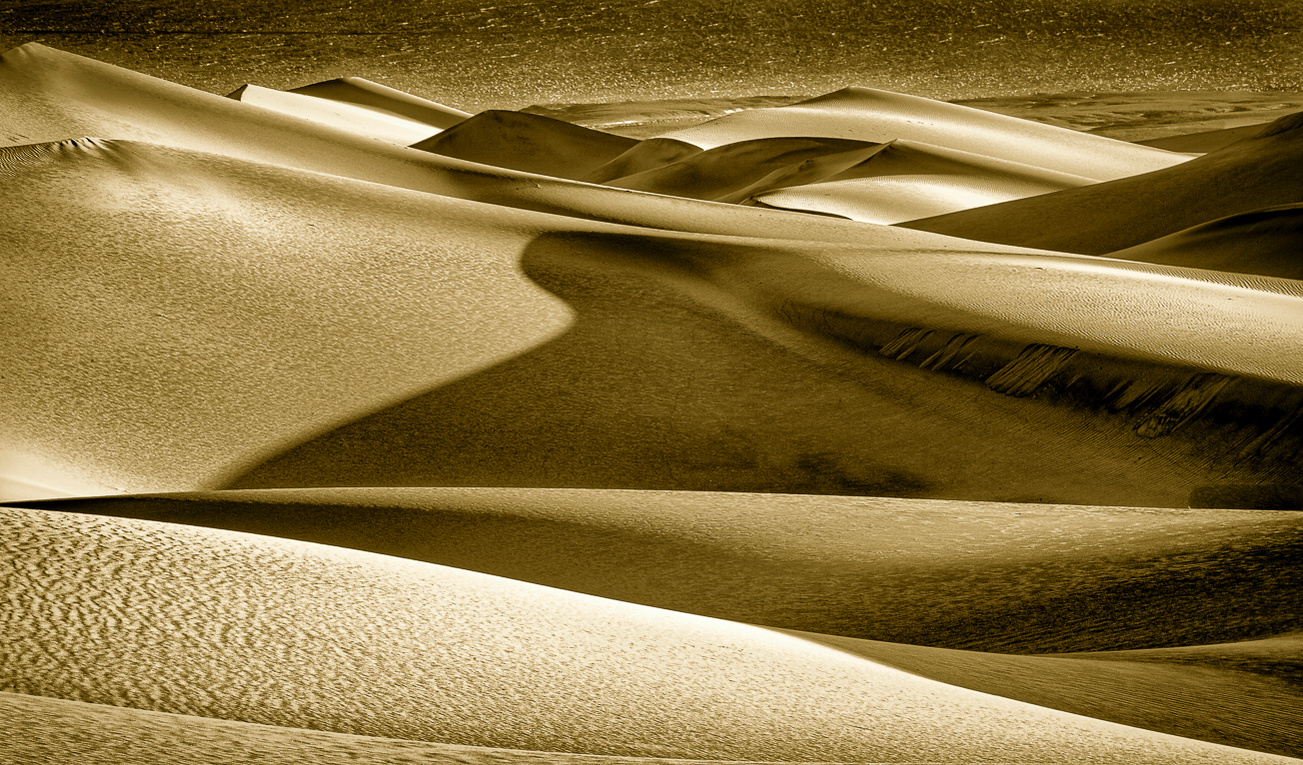 A warm-toned black and white image of a Death Valley sand dune highlighting the dune's contours, shape, texture, shadows, and fine details.