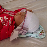 Head and shoulders/torso photo of a baby lying on its back supine on a bed wearing fuchsia pink with a pale pink head scarf.  The baby is asleep with its right hand by its right cheek.