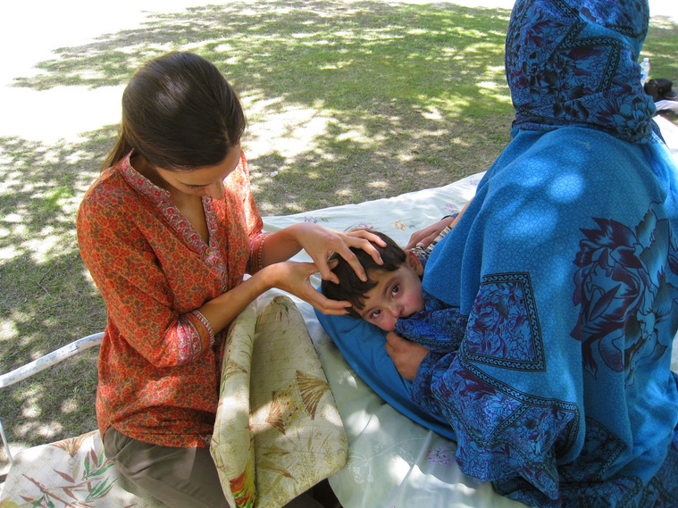 Katy kneels alongside a charpoy on which a local Pakistani lady sits cross-legged holding her baby who Katy is treating with cranial osteopathy; using a highly trained sense of touch to feel subtle changes of tension and tissue quality. Nagar Valley.