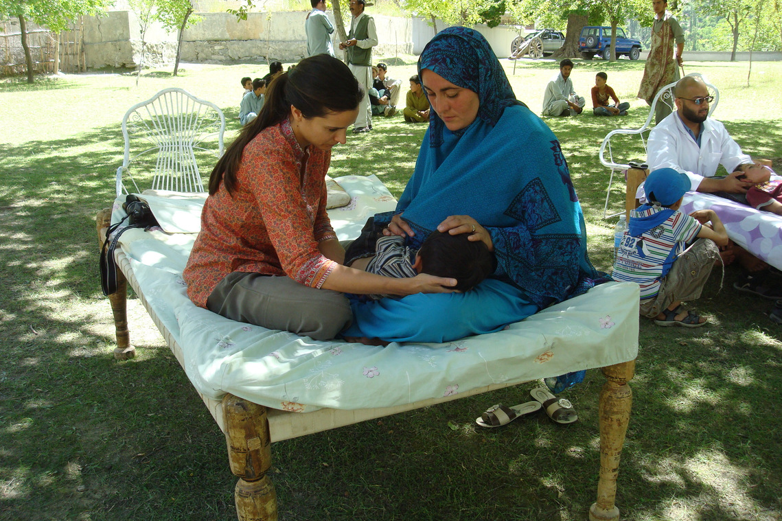 Katy is sitting cross-legged on a charpoy, with a Pakistani lady who is wearing a turquoise burqa/sari.  This lady's baby is laid across both laps whilst being treated with cranial osteopathy.  Faisal Naqvi practices alongside and Nagar villagers look on.