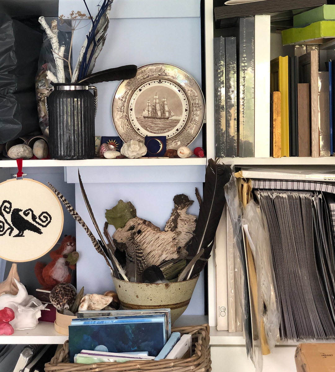 A close up of some studio shelves containing coloured twigs, a sepia coloured vintage plate with a frigate on it, a bowl of bark and feathers, a cross stitched crow on a loop and frames and canvases. 