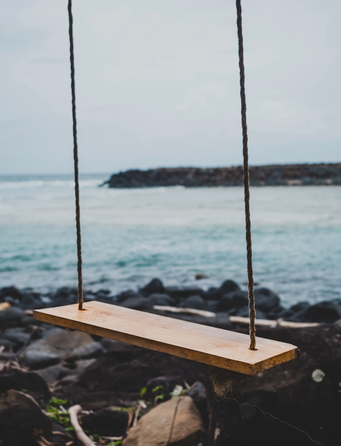 An empty wooden swing, hanging on a rocky beach waiting for a sitter.