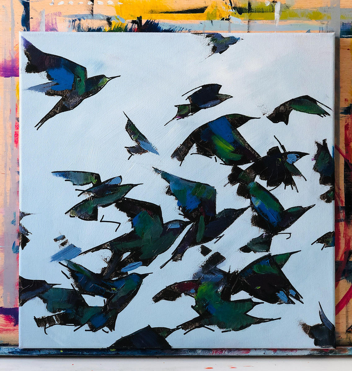 A canvas against a painting board.  The canvas contains a flock of starlings flying up bottom left to top right, they are black and green and blue.  Their wings are mid beat, all in different stages of uplift.