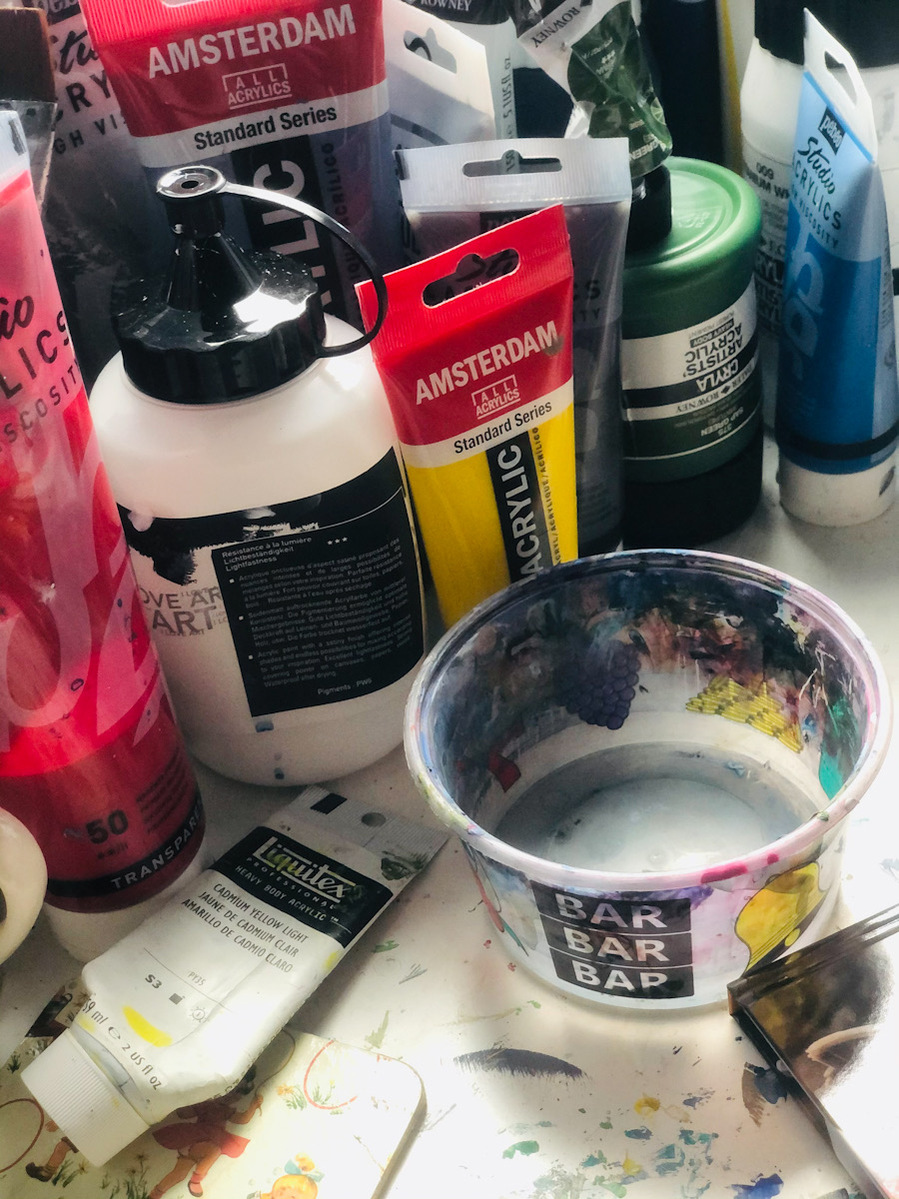 Acrylic paints (Amsterdam, I love art, Daler Rowney Cryla and Pebeo brands) and a messy table below, a slot machine plastic pot is covered in paint, it's used to hold paint water. 