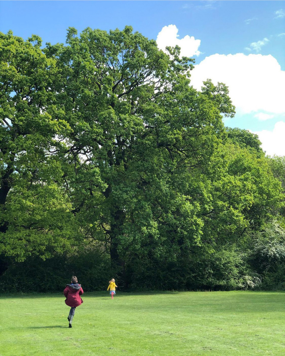 Two children running across a large green lawn into the distant verdant treeline, coats flapping in the wind.  The sky is blue with white clouds.