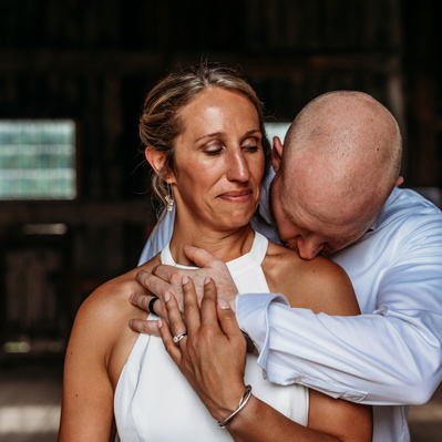 The bride and groom stand together in a sunlit field, their faces glowing with joy and love. The bride, in her elegant white gown, holds a bouquet of soft pastel flowers, while her veil flows gently in the breeze. The groom, in a classic black tuxedo, sta