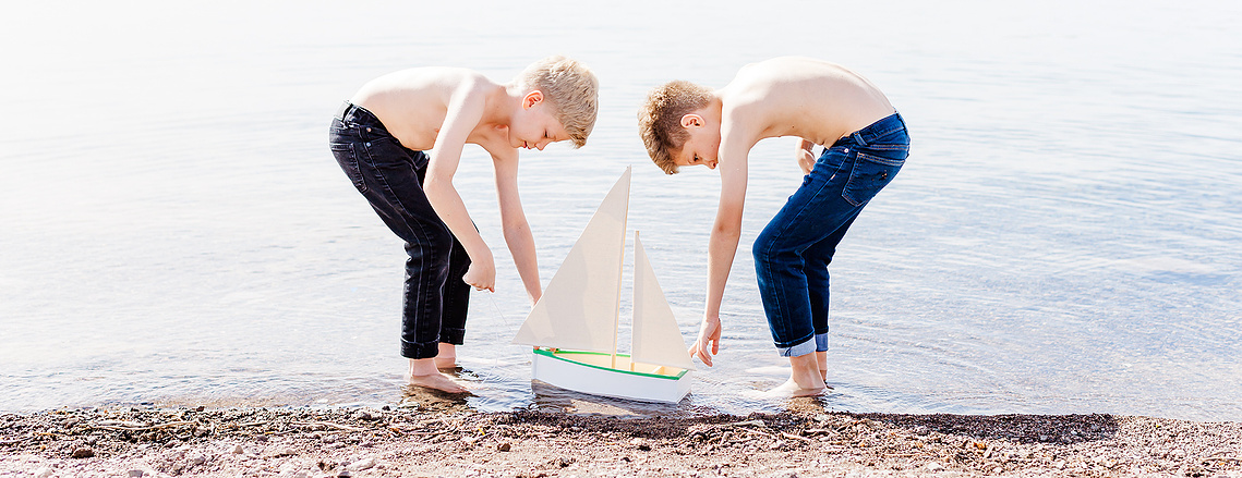 Two boys playing at the water's edge with a small sailboat.