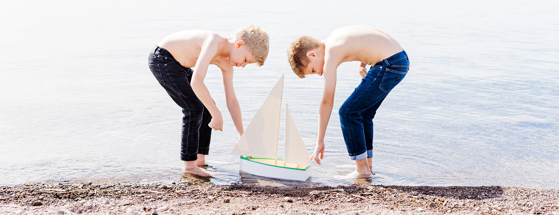 Two boys playing at the water's edge with a small sailboat.