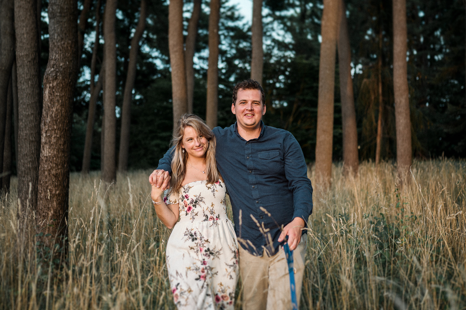 a couple walks with their arms around each other through tall grass and trees in the arnold arboretum