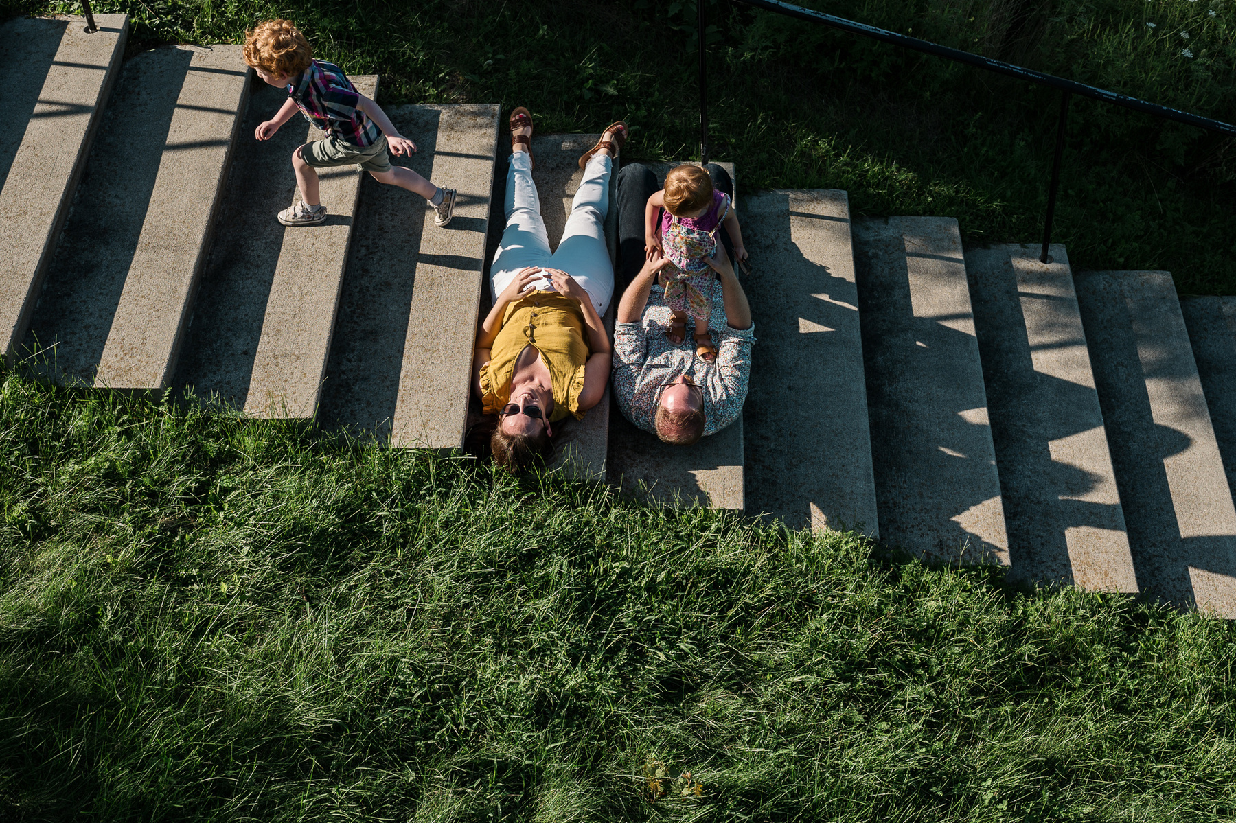 A couple with sunglasses on lays in the light while their kids play near them on the stairs at Fisher Hill Reservoir Park in Chestnut Hill by Brookline family photographer Korri Leigh Photography