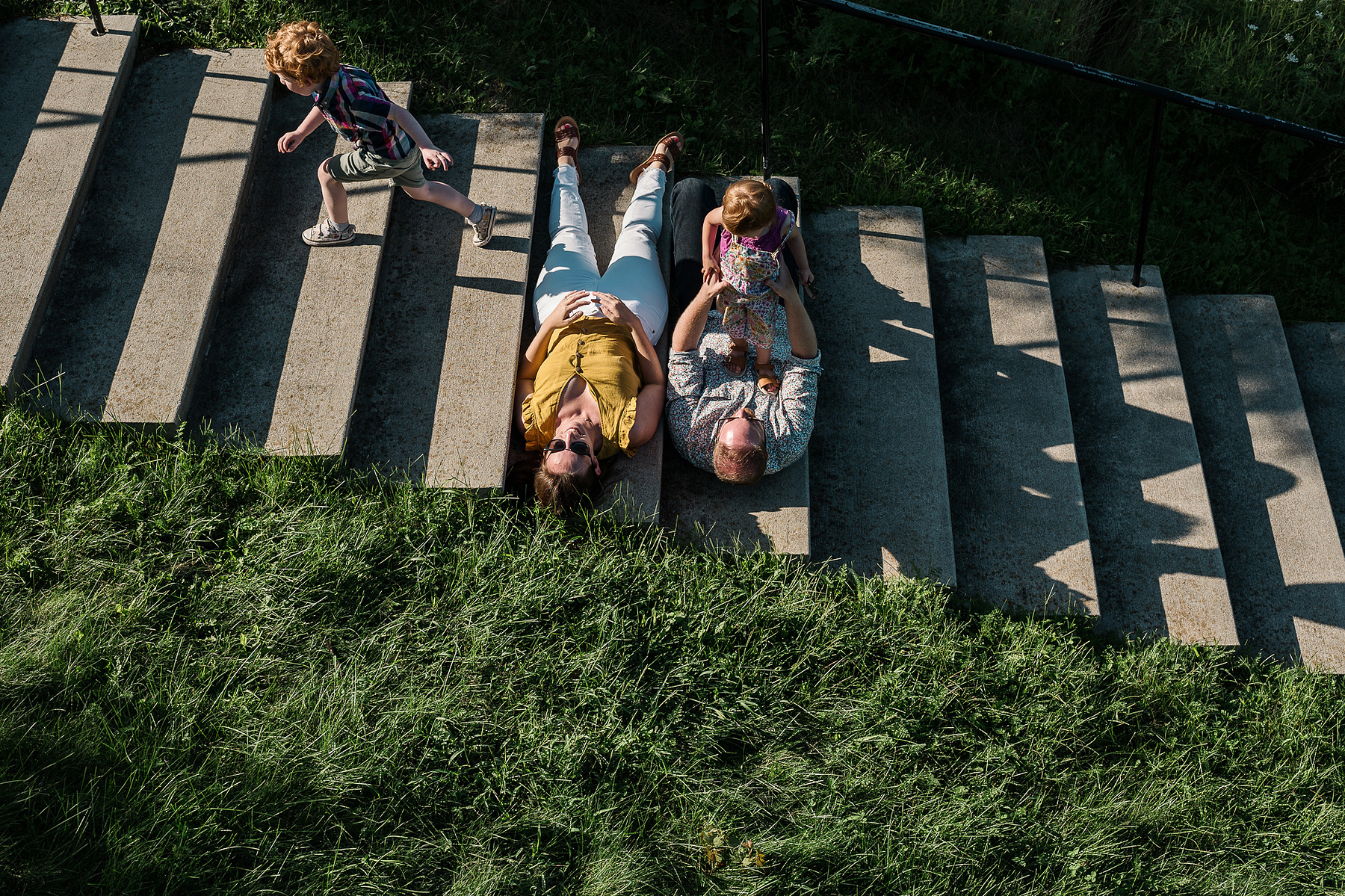 A couple with sunglasses on lays in the light while their kids play near them on the stairs at Fisher Hill Reservoir Park in Chestnut Hill by Brookline family photographer Korri Leigh Photography