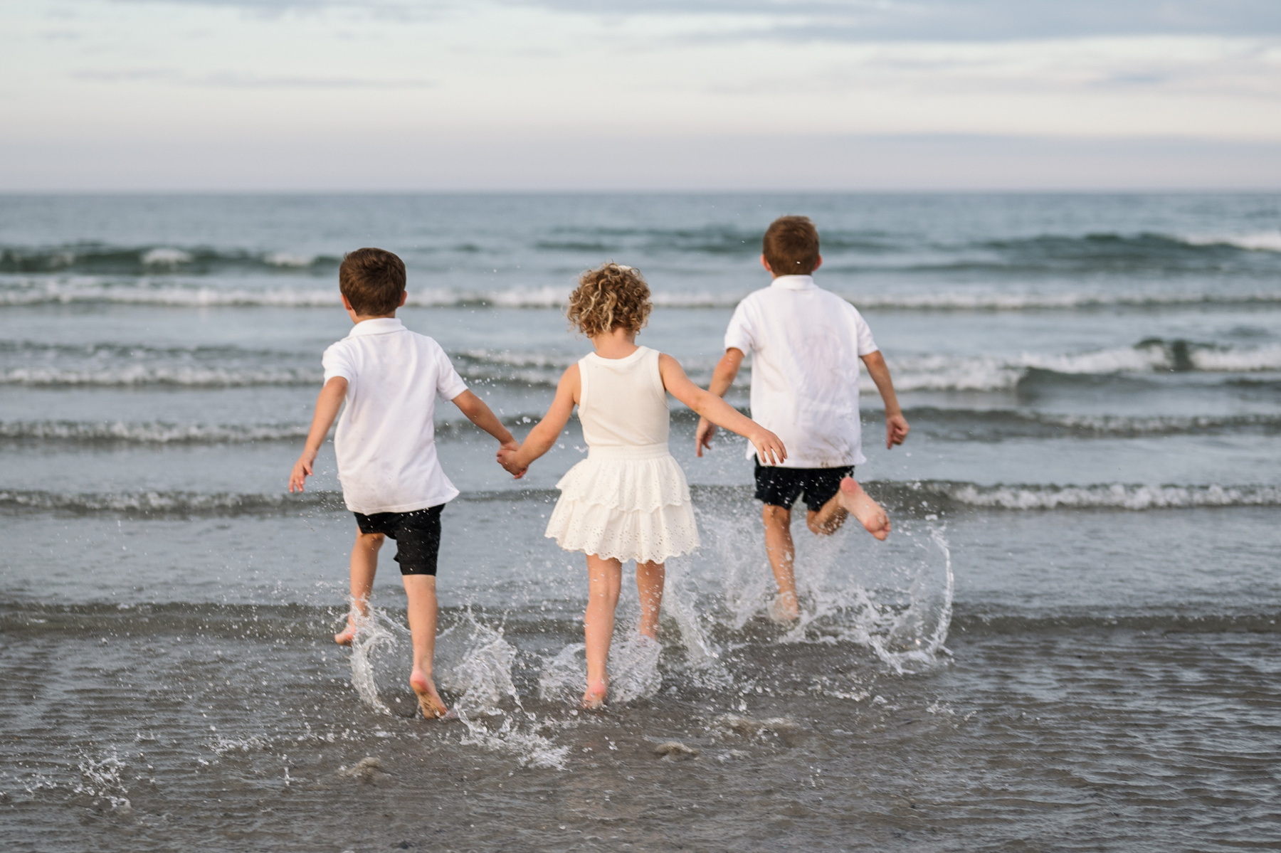 Three children hold hands and run into the Atlantic Ocean at Nantasket Beach in Hull during their summer vacation photography session by Boston Family Photographer Korri Leigh Crowley