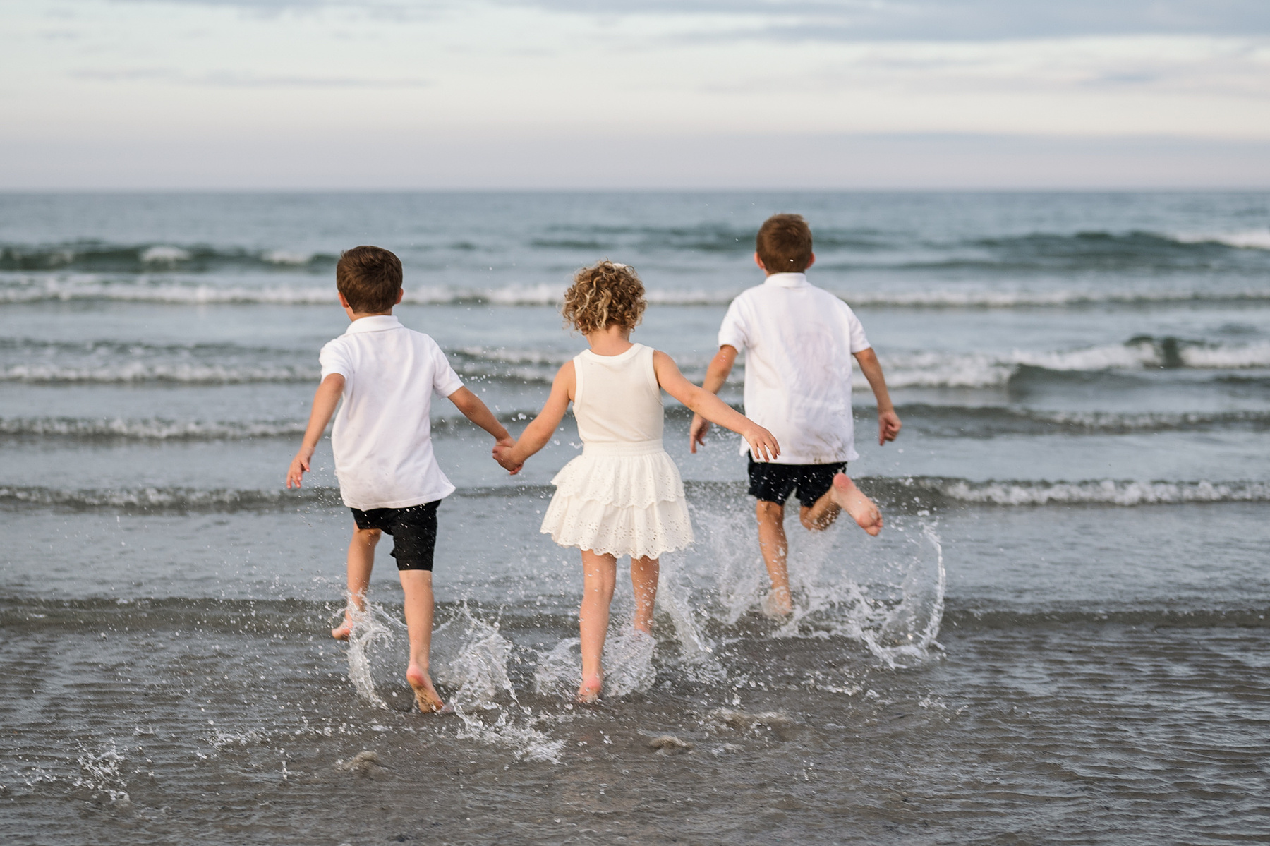 Three children hold hands and run into the Atlantic Ocean at Nantasket Beach in Hull during their summer vacation photography session by Boston Family Photographer Korri Leigh Crowley