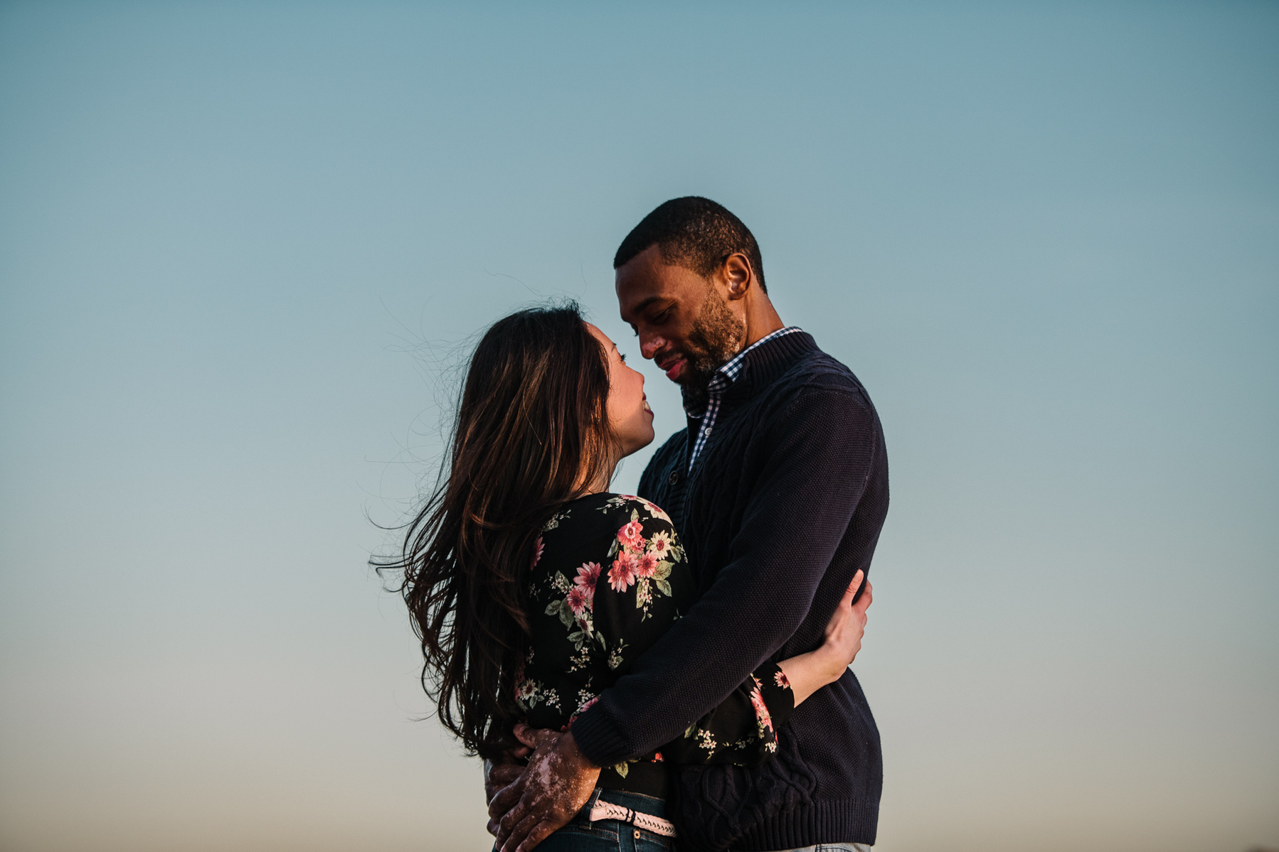 A couples stands with their arms around each other in front on a colorful sky in charlestown massachusetts