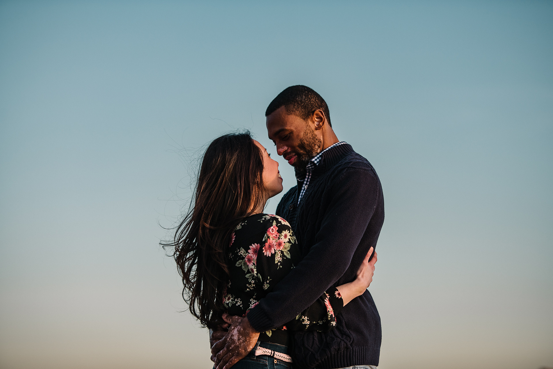 A couples stands with their arms around each other in front on a colorful sky in charlestown massachusetts
