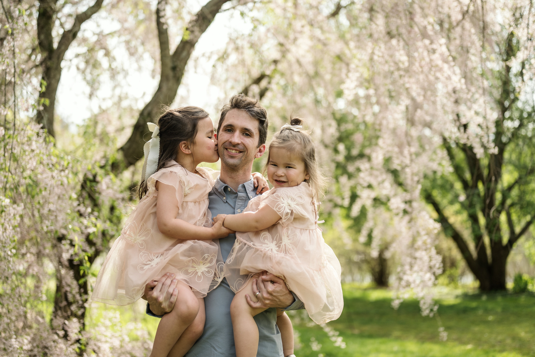 Two girls snuggle with their father for a photo in front of pink flowering trees in Boston's Arnold Arboretum in spring.  Photo by West Roxbury family photographer Korri Leigh Photography