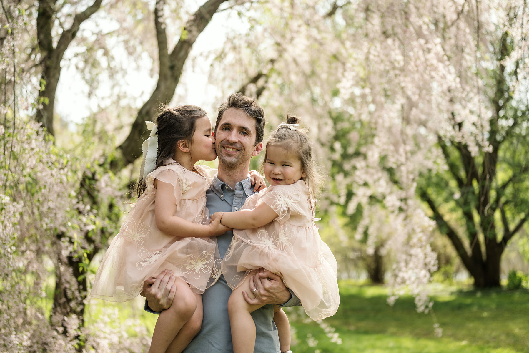 Two girls snuggle with their father for a photo in front of pink flowering trees in Boston's Arnold Arboretum in spring.  Photo by West Roxbury family photographer Korri Leigh Photography