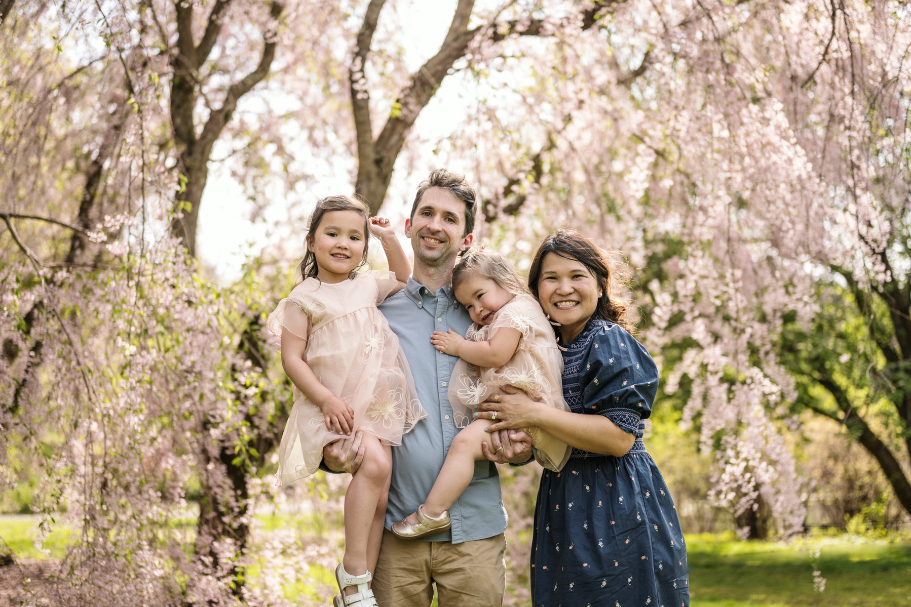 A family smiles for a photo in front of pink flowering trees in Boston's Arnold Arboretum in spring.  Photo by West Roxbury family photographer Korri Leigh Photography
