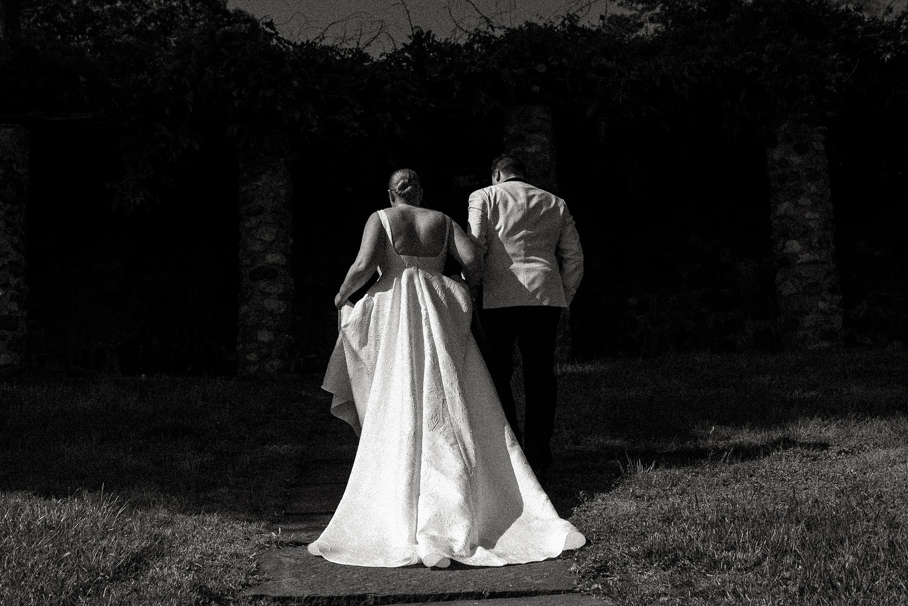 Viewed from behind, a bride and groom walk hand and hand into a shadow at The Crane Estate in Ipswich MA