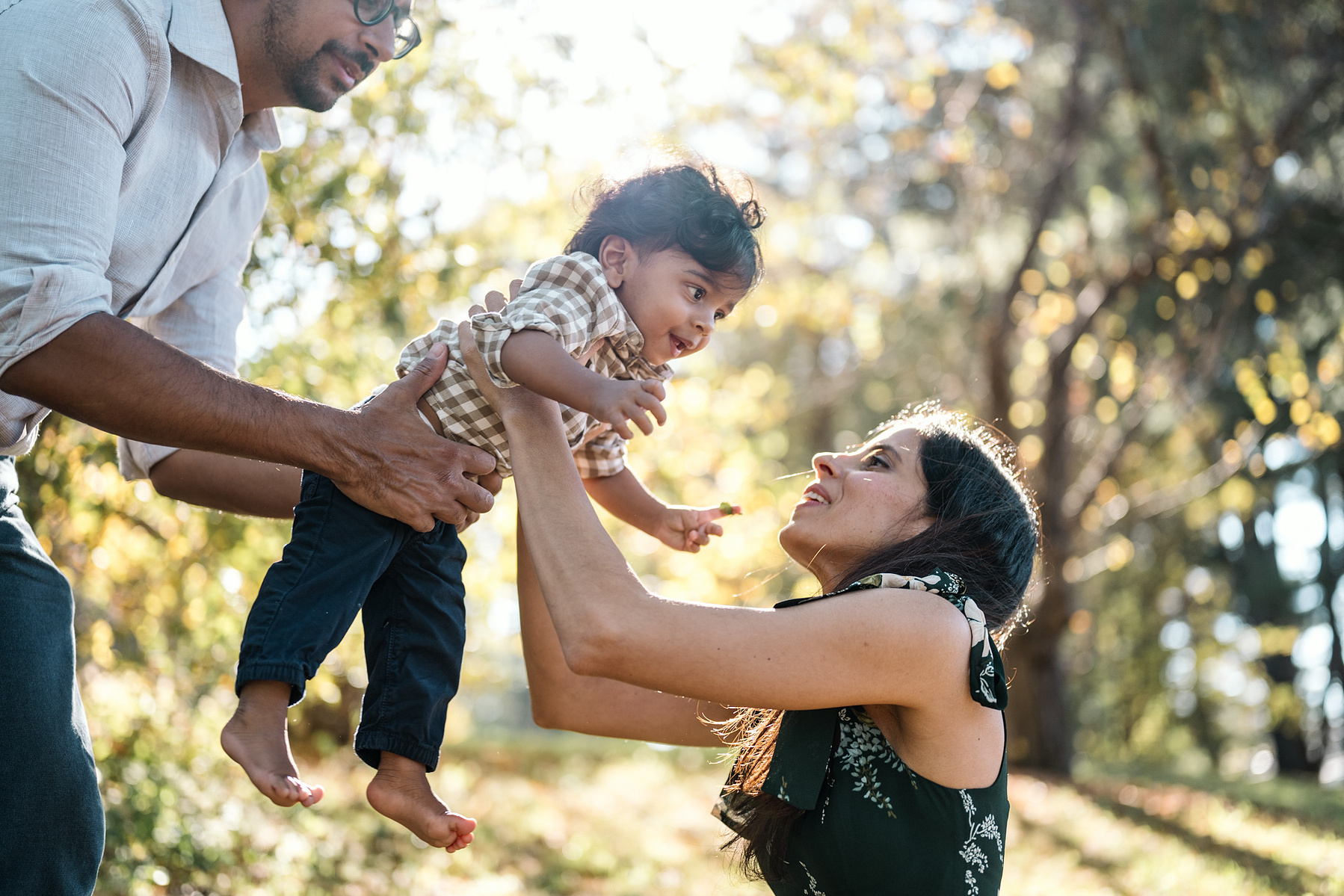A baby is passed between parents in the arnold arboretum during their boston family photography session by west roxbury family photographer korri leigh crowley