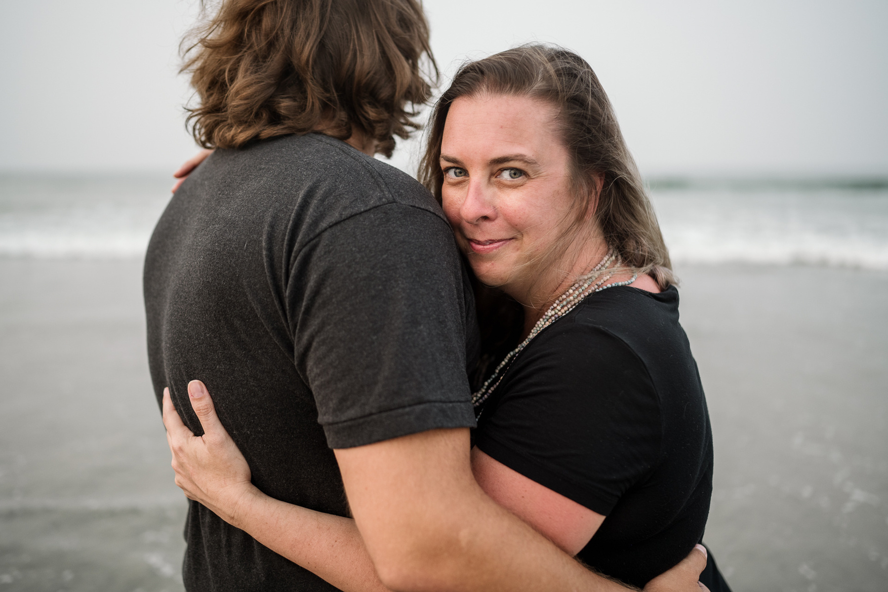 A woman peeks over her partner's shoulder on Nantasket Beach at their Hull family photography session by Korri Leigh Photography
