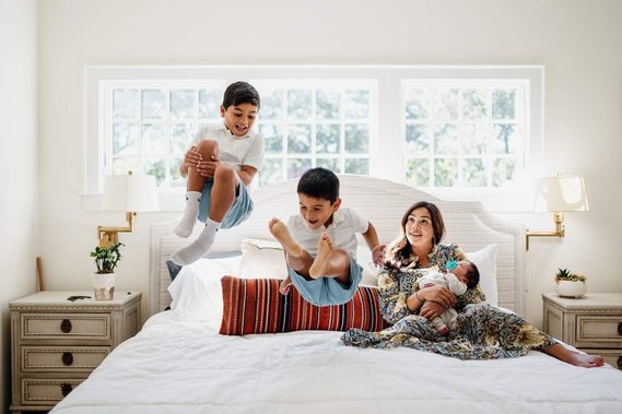 Two young boys jump on a large bed while their mother feeds a baby and looks on with a smile during a Hingham family photoshoot 
