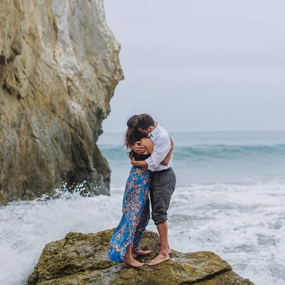 A couple embraces on a rock in the ocean  during their El Matador Beach engagement session