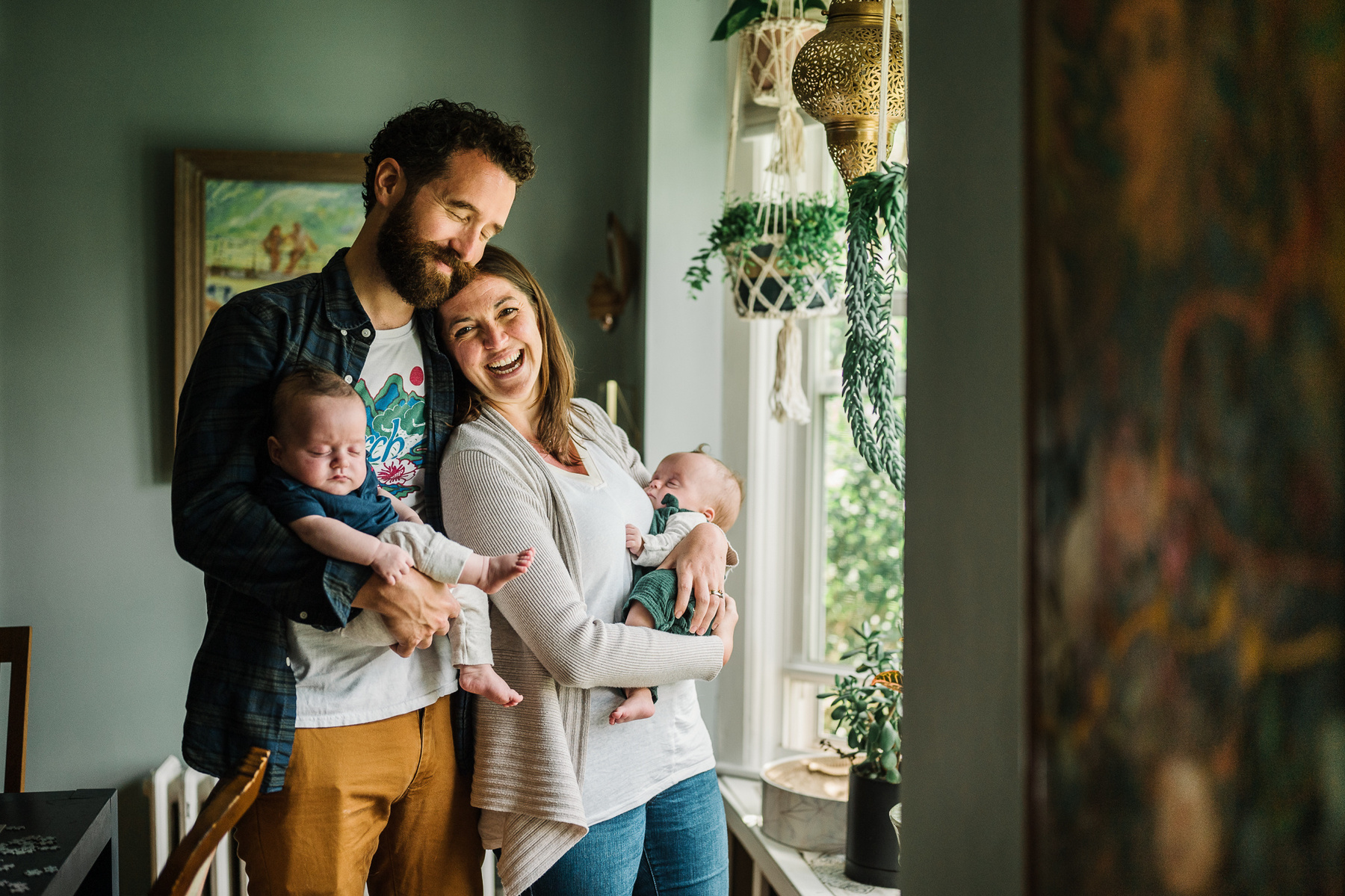 Parents stand in a window holding their newborn twins and snuggle and smile for the camera
