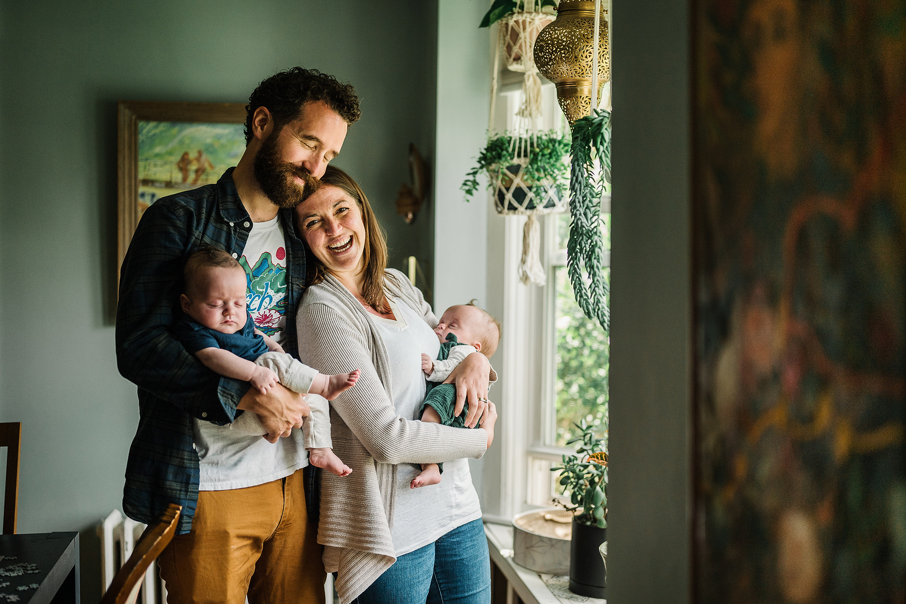 Parents stand in a window holding their newborn twins and snuggle and smile for the camera