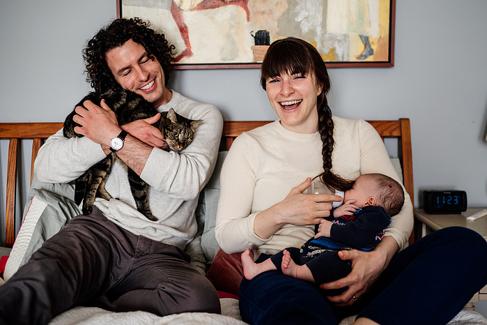 A mother feeds a baby a bottle while her husband cuddles the cat while they both sit on a bed and laugh