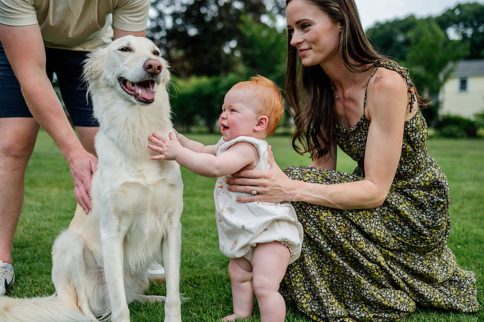 A mother in a long floral green dress holds her daughter who pets a medium white long haired dog