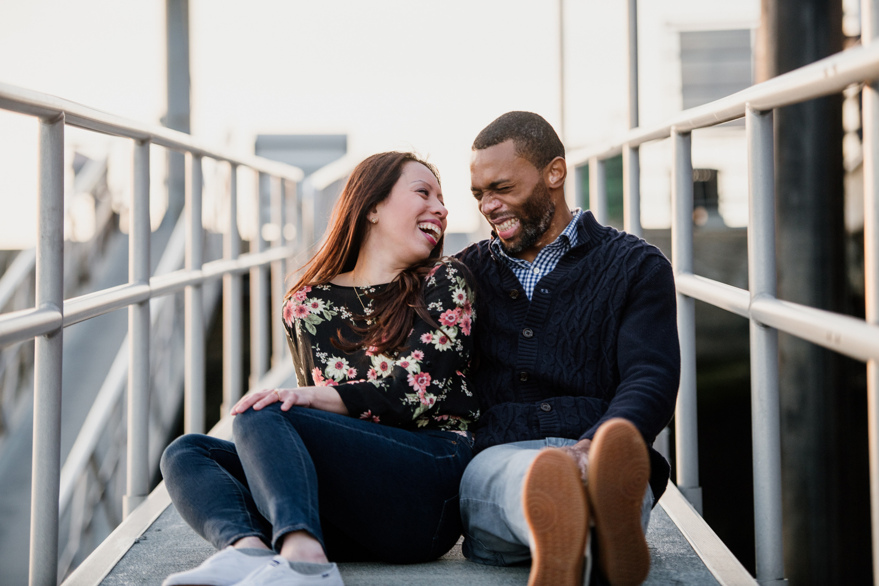 A couples sits on a dock ramp in charlestown while looking at each other and laughing after their boston proposal