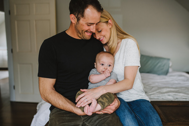 A father in a black t-shirt sits on a bed while his wife in a white t-shirt cuddles him and their young son, held on their laps