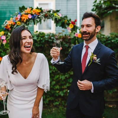 An attractive couple laughs while holding champagne glasses during their cambridge backyard microwedding