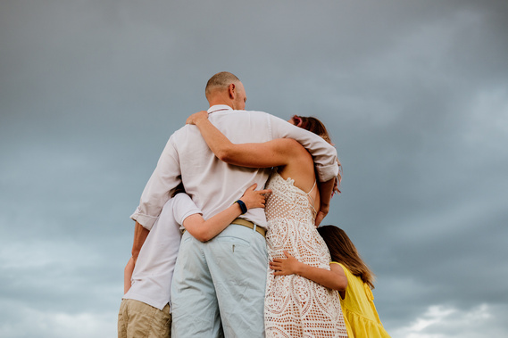 A family of four is shown from behind against a cloudy sky, their arms around each other's backs