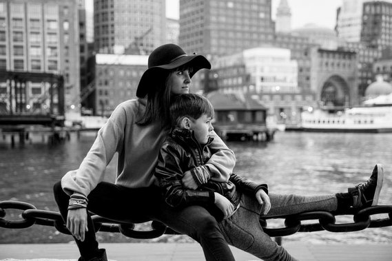 Boston photographer Korri Leigh Crowley sits on a large chain fence with her son in front of the Boston skyline