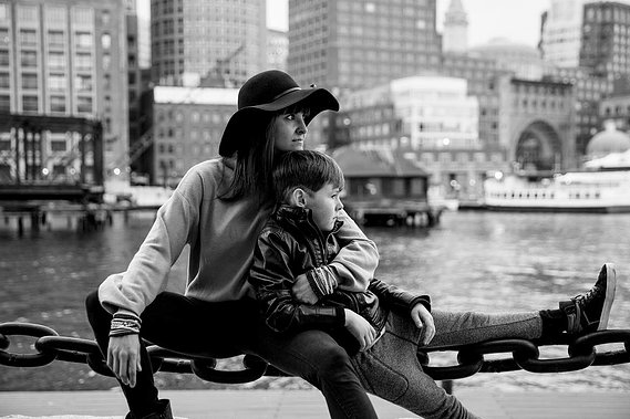Boston photographer Korri Leigh Crowley sits on a large chain fence with her son in front of the Boston skyline