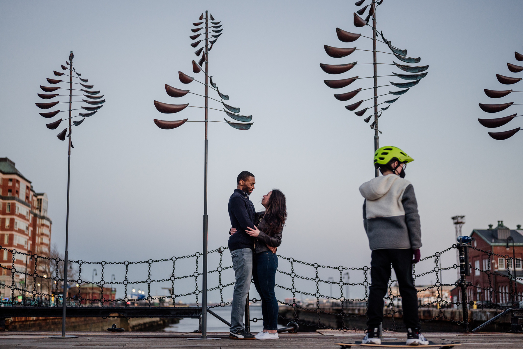 A couple stands in front of a large public art piece in the charlestown navy yard while a young boy skateboards by in the foreground