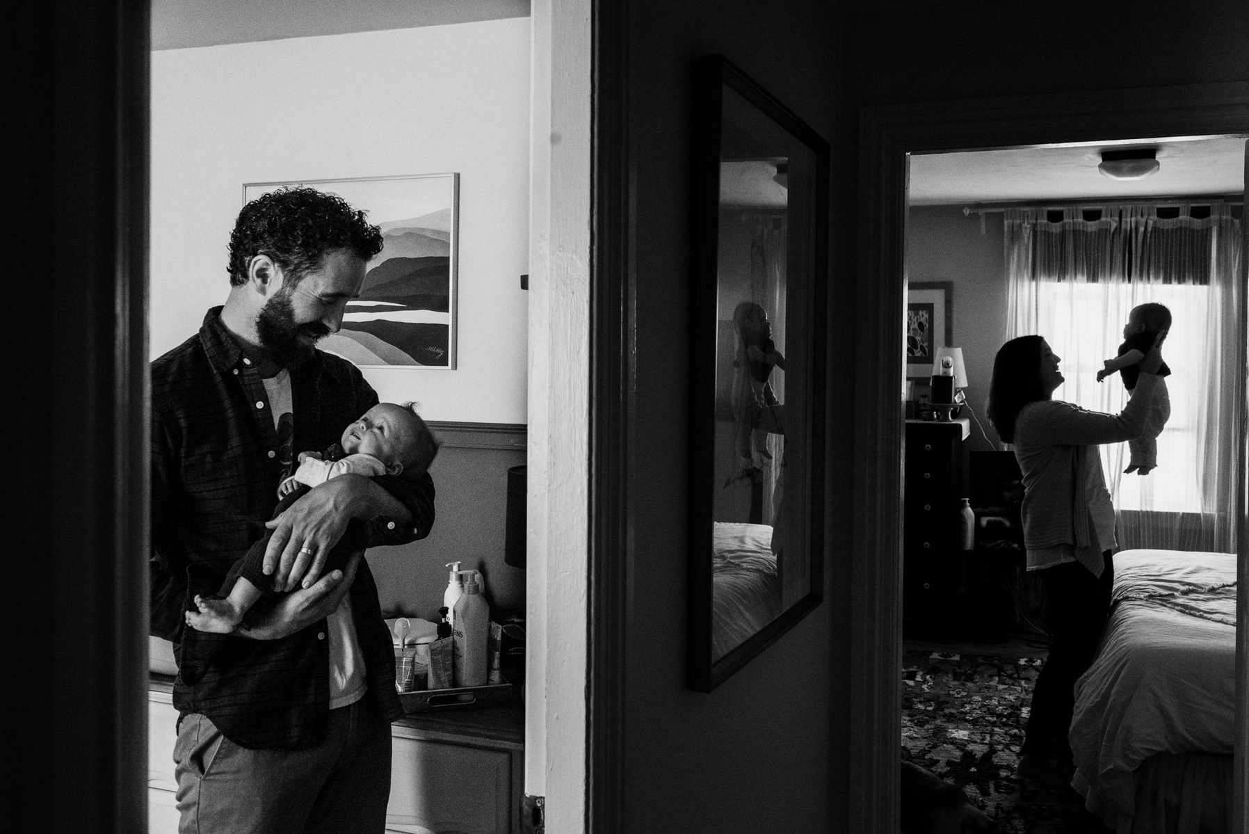 A father holds their newborn twin daughter in a doorway while his wife holds their newborn son in another room down the hall