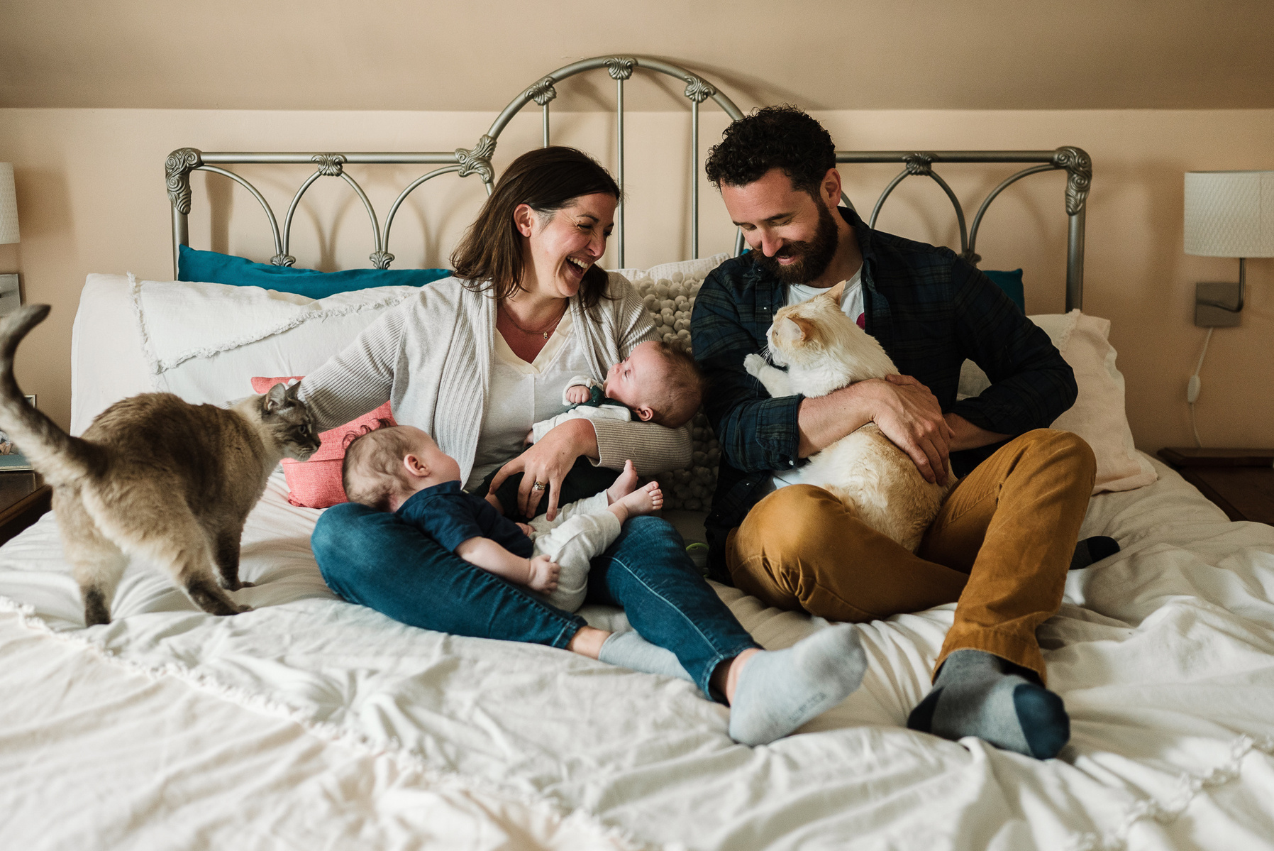 Parents snuggle their newborn twins while reclining on their bed and playing with their cats