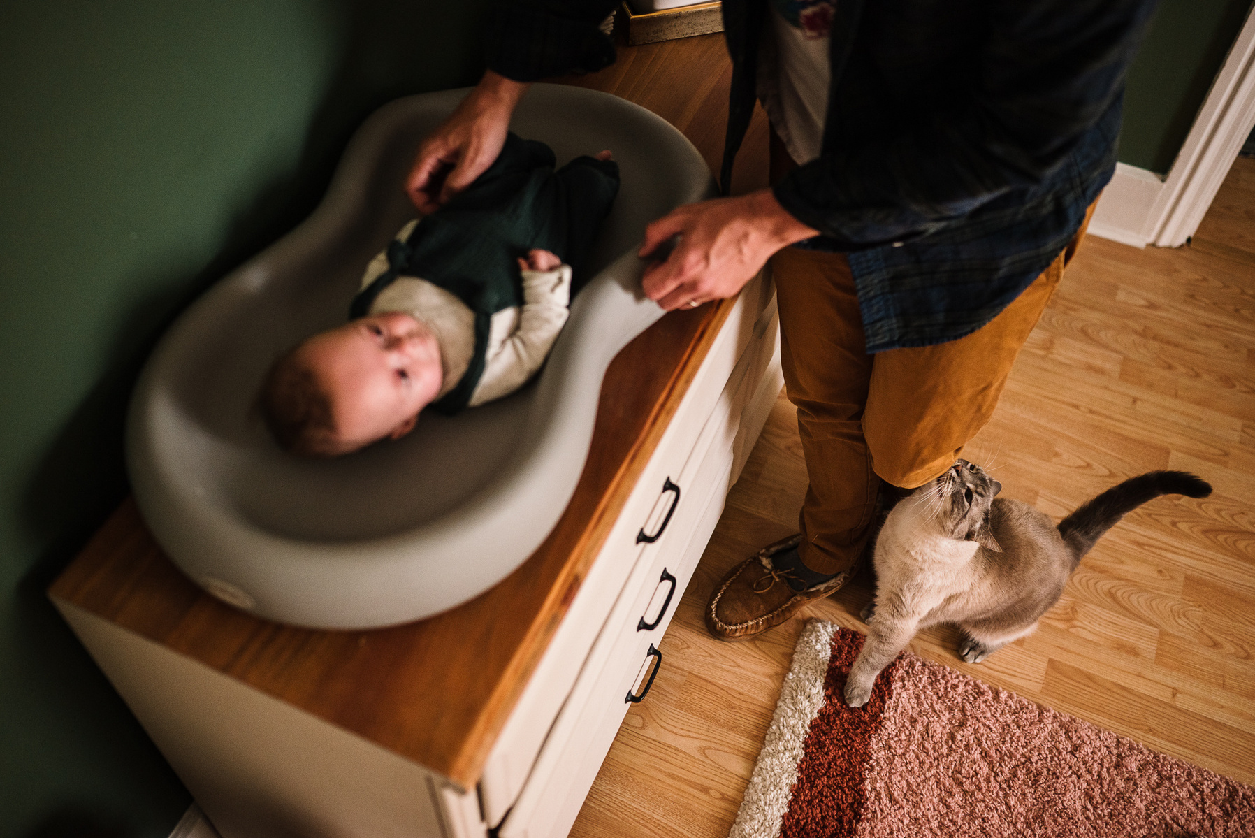 A newborn twin lays on a changing table while a cat rubs on her father's legs