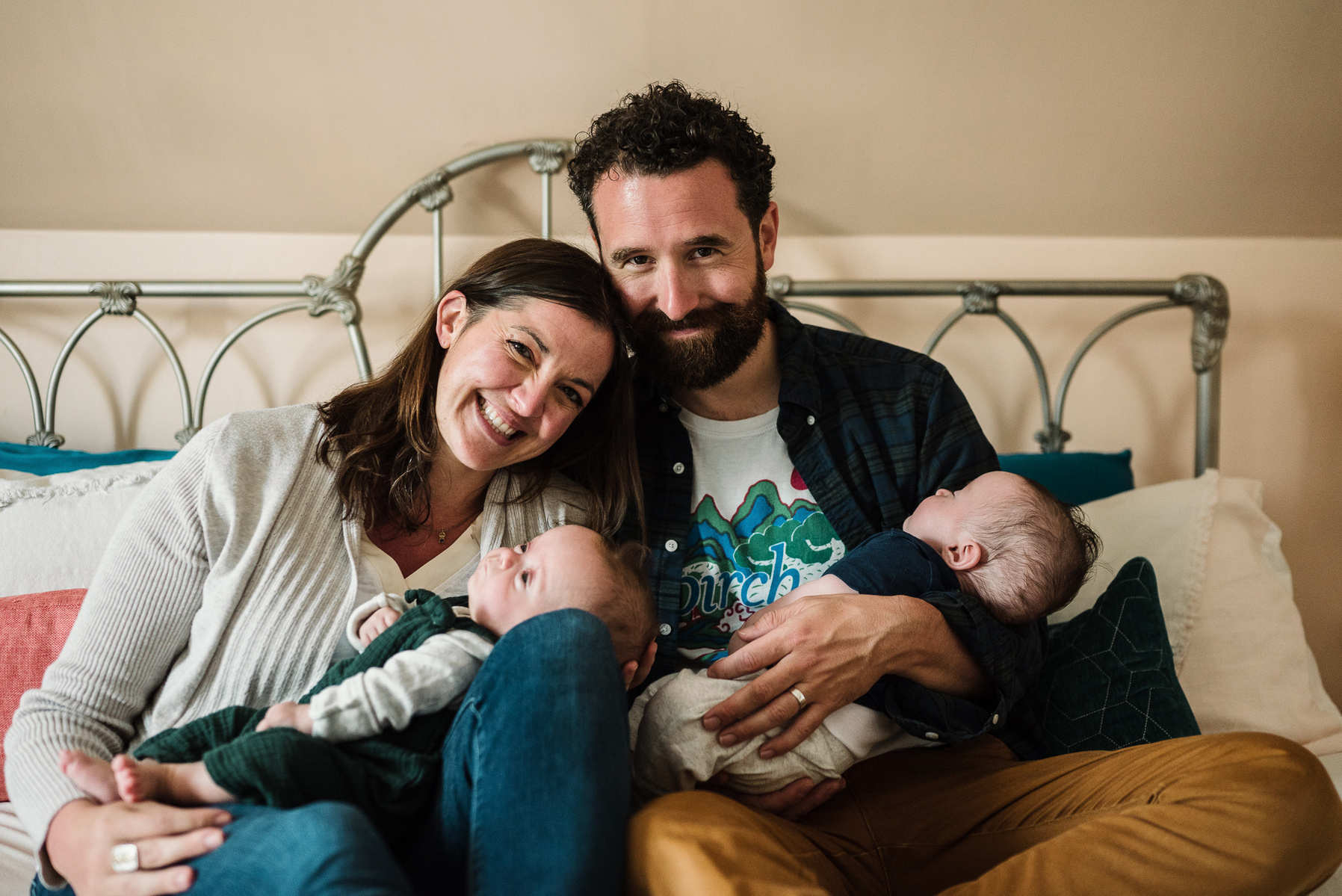 Parents snuggle their newborn twins while reclining on their bed and smiling at the camera