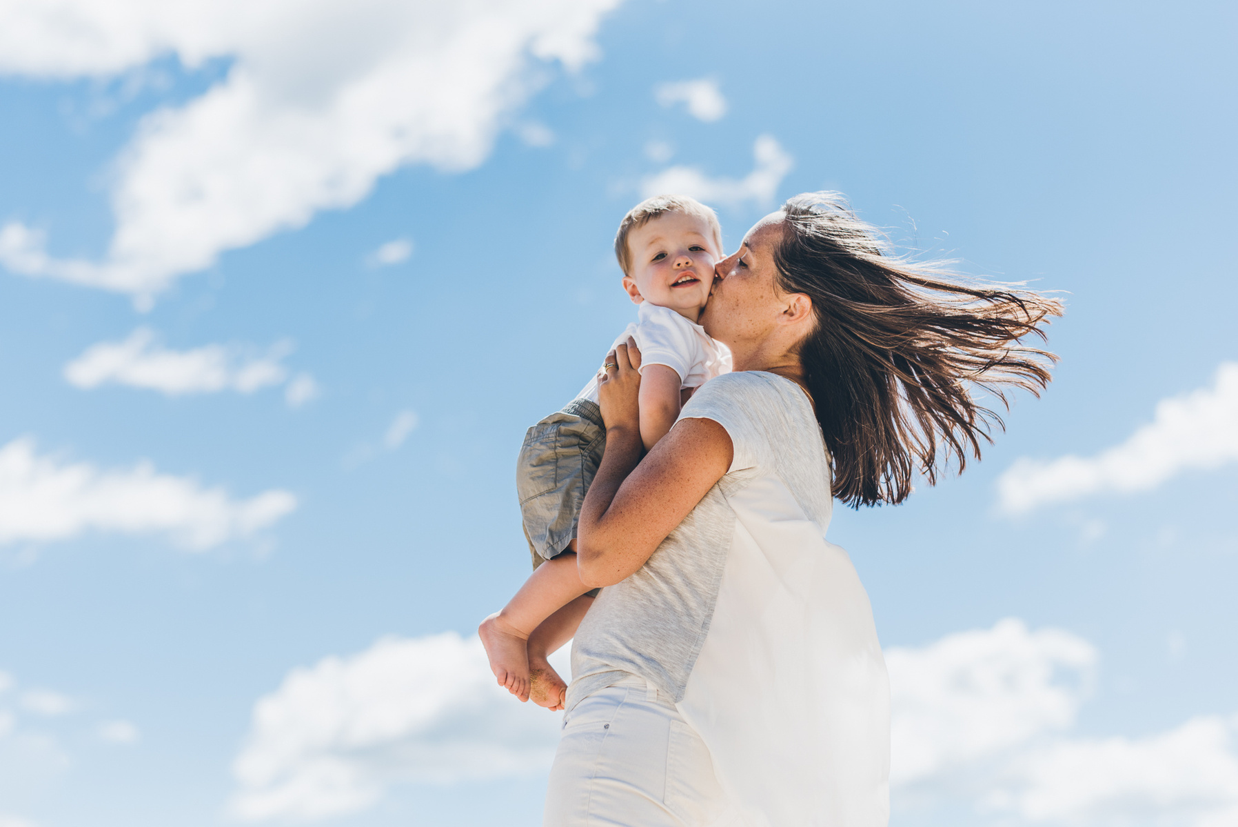 A mother lifts her son into the air and kisses his cheek while framed against a blue summer sky during their cape cod family photography session