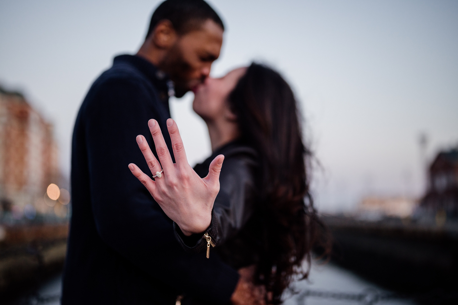 a woman shows off her engagement ring in the foreground while she kisses her fiancee in the background