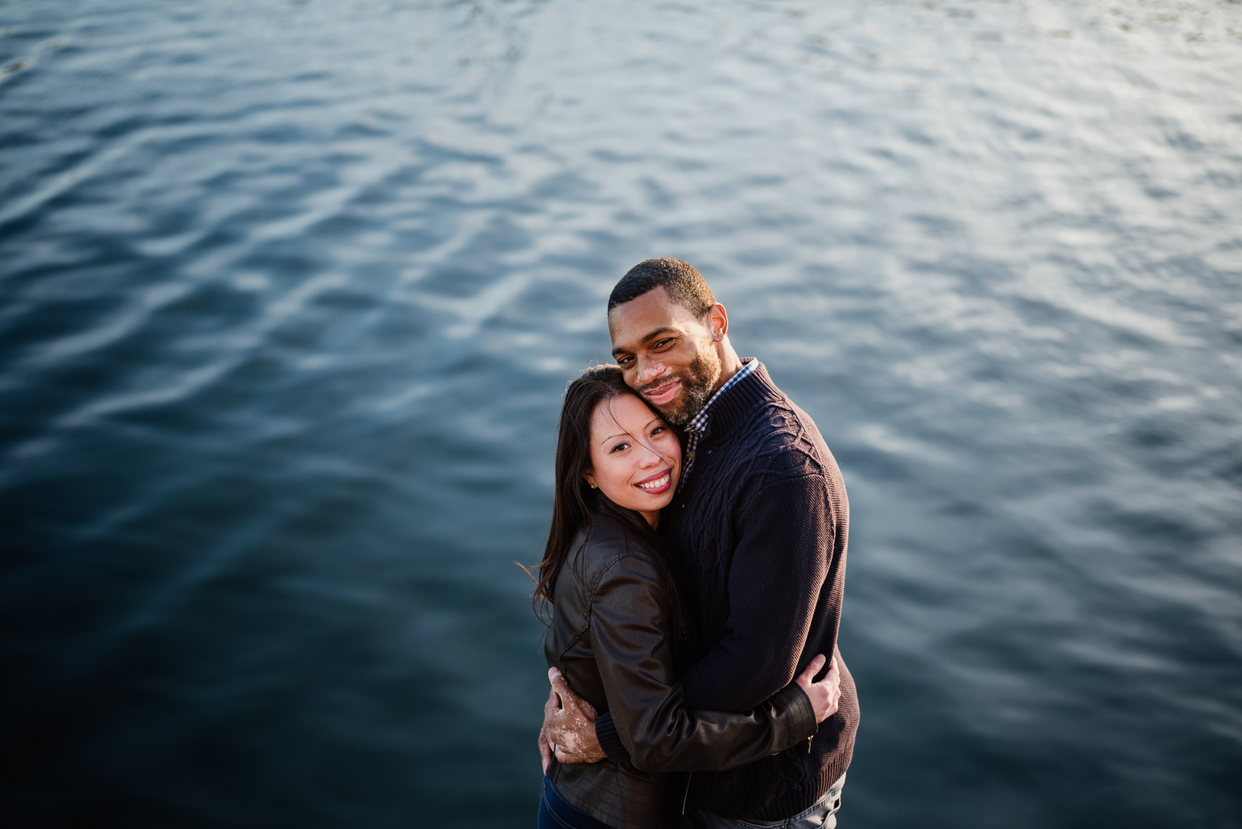 a couple snuggles and looks at the camera while standing in front of boston harbor rippled with small waves