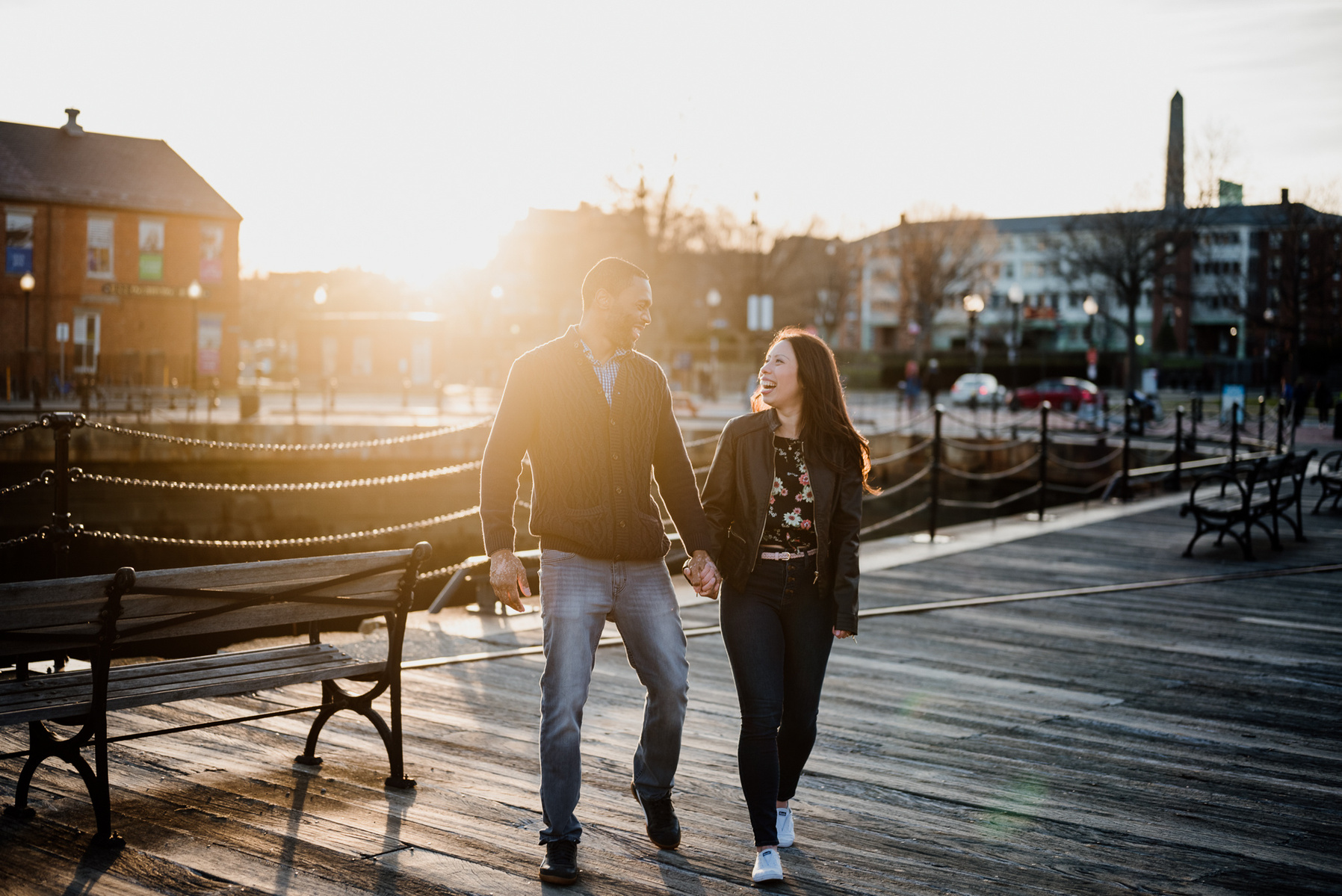 A couples holds hands and walks in front maritime buildings at sunset