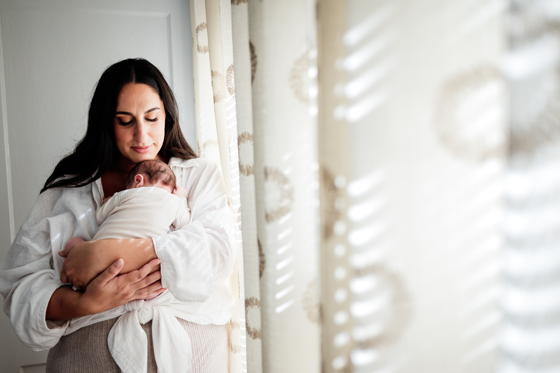 a mother cuddles her baby under her chin while standing by white sun-lit curtains during her boston newborn photography session