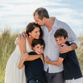 A family of 4 hugs and smiles for the camera on the beach on a sunny day