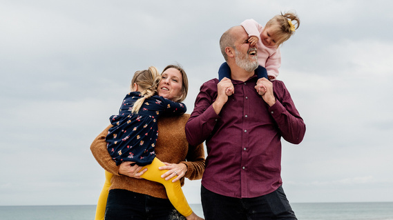 A toddler on her father's shoulder grabs his nose while the mother hugs their other child.  The family of four smiles and play on an overcast beach.